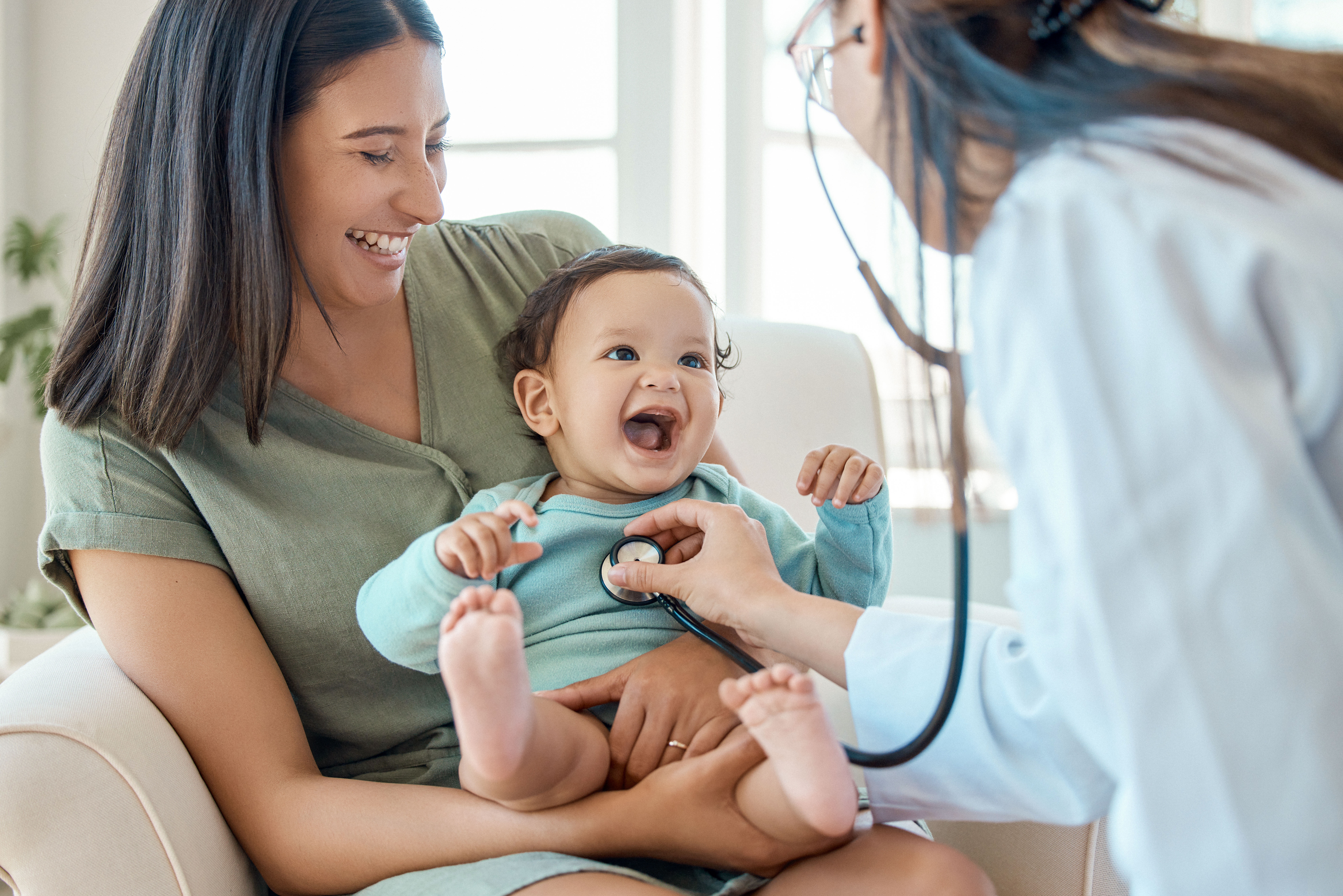 Baby sitzt auf dem Schoß der Mutter und wird von einer Krankenschwester mit einem Stethoskop untersucht